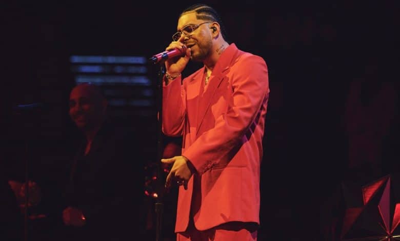 A male singer performs on stage in a vibrant red suit and glasses, holding a microphone. soft, warm lighting illuminates him and a blurred figure in the background, likely a band member.