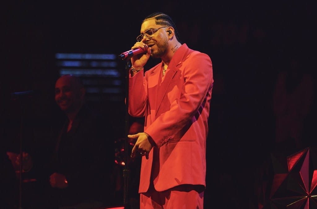 A male singer performs on stage in a vibrant red suit and glasses, holding a microphone. soft, warm lighting illuminates him and a blurred figure in the background, likely a band member.