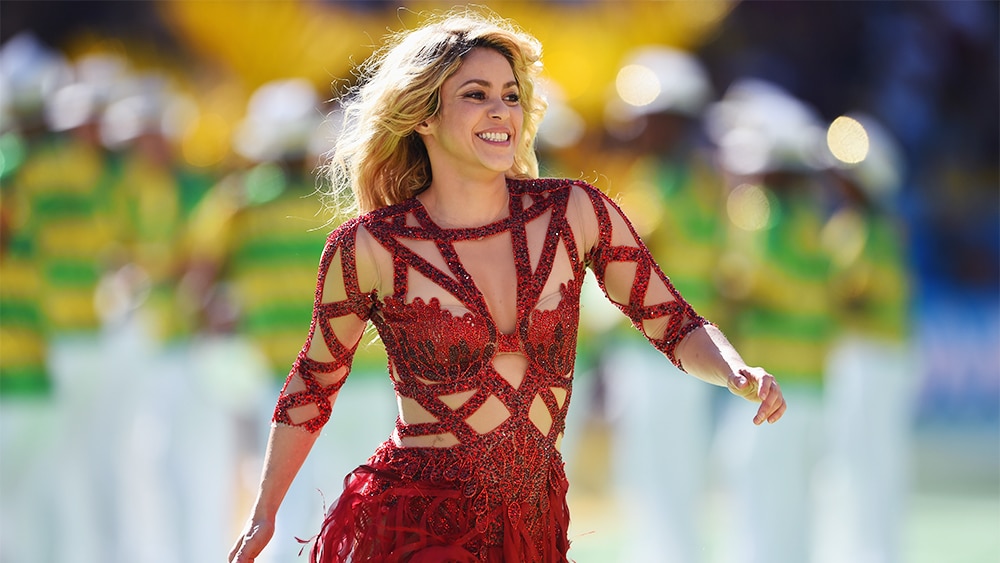 A woman in a sparkling red dress performs on a sunlit stage during Shakira's 'Las Mujeres Ya No Lloran' World Tour, her joyful expression and dynamic pose capturing the celebratory