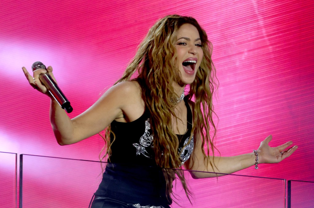 A female singer with long, wavy blonde hair energetically performs Shakira's 'Waka Waka' on stage against a bright pink background. She wears a black top with silver embellishments