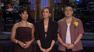 Three people stand side by side on a stage with musical equipment in the background. From left to right: Kristen Wiig in a brown dress, a woman in a black blazer, and a man