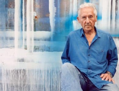An elderly man with gray hair sits in front of a vibrant blue and white abstract painted wall, reminiscent of Ed Ruscha's style. He is wearing a blue shirt and jeans, looking calmly at the