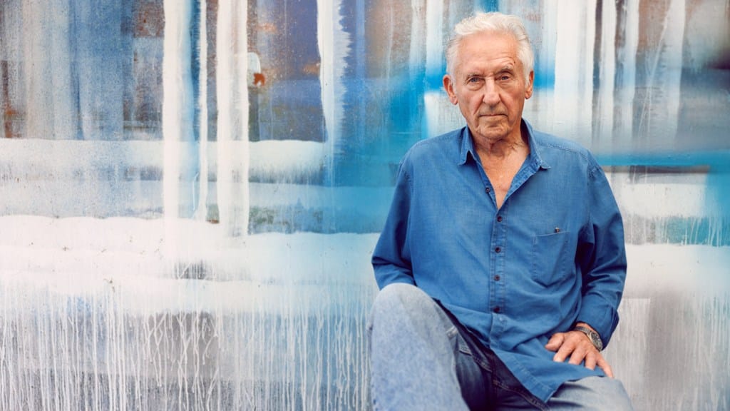An elderly man with gray hair sits in front of a vibrant blue and white abstract painted wall, reminiscent of Ed Ruscha's style. He is wearing a blue shirt and jeans, looking calmly at the