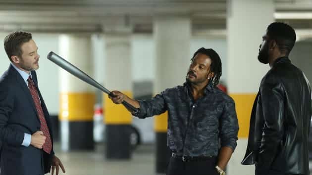 Three men in a parking garage engaging tensely. One man in a suit faces a man with long hair who points a large metal tube at him, while another man, resembling the complex villain Elijah Stone