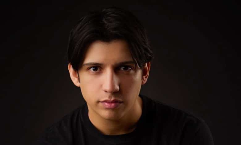 A portrait of Kevin Alves, a young man with medium-length dark hair and fair skin, wearing a black t-shirt. He has a serious expression, looking directly at the camera, set against a