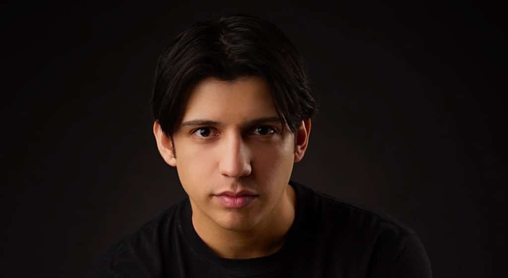 A portrait of Kevin Alves, a young man with medium-length dark hair and fair skin, wearing a black t-shirt. He has a serious expression, looking directly at the camera, set against a