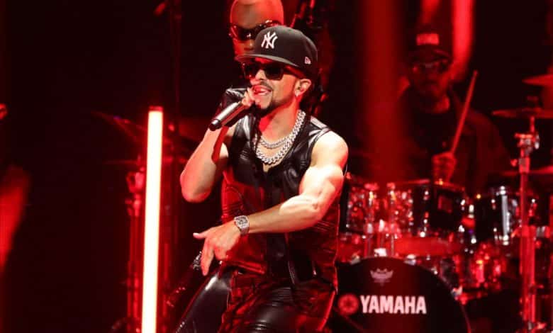 A male singer performs onstage, wearing a black tank top, sunglasses, and a ny baseball cap. he's surrounded by red stage lights with a drummer visible in the background, near a yamaha drum set.