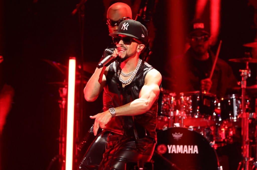 A male singer performs onstage, wearing a black tank top, sunglasses, and a ny baseball cap. he's surrounded by red stage lights with a drummer visible in the background, near a yamaha drum set.