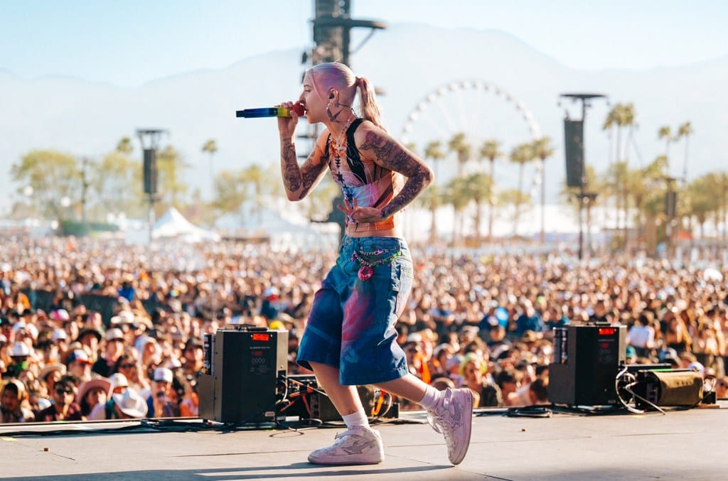 A young woman with vibrant pink hair and tattoos performs energetically on stage at Coachella, holding a microphone. She's dressed in colorful denim, facing a large, sunlit crowd with a Ferr