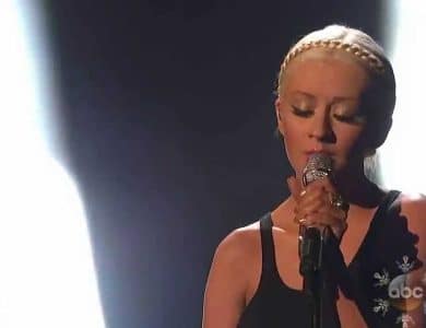 A woman with blonde hair styled in braids sings into a microphone, spotlighted on a dimly lit stage, during her emotive "Say Something" performance. Her expression is focused and intense.