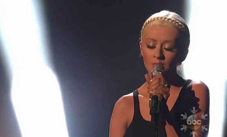 A woman with blonde hair styled in braids sings into a microphone, spotlighted on a dimly lit stage, during her emotive "Say Something" performance. Her expression is focused and intense.