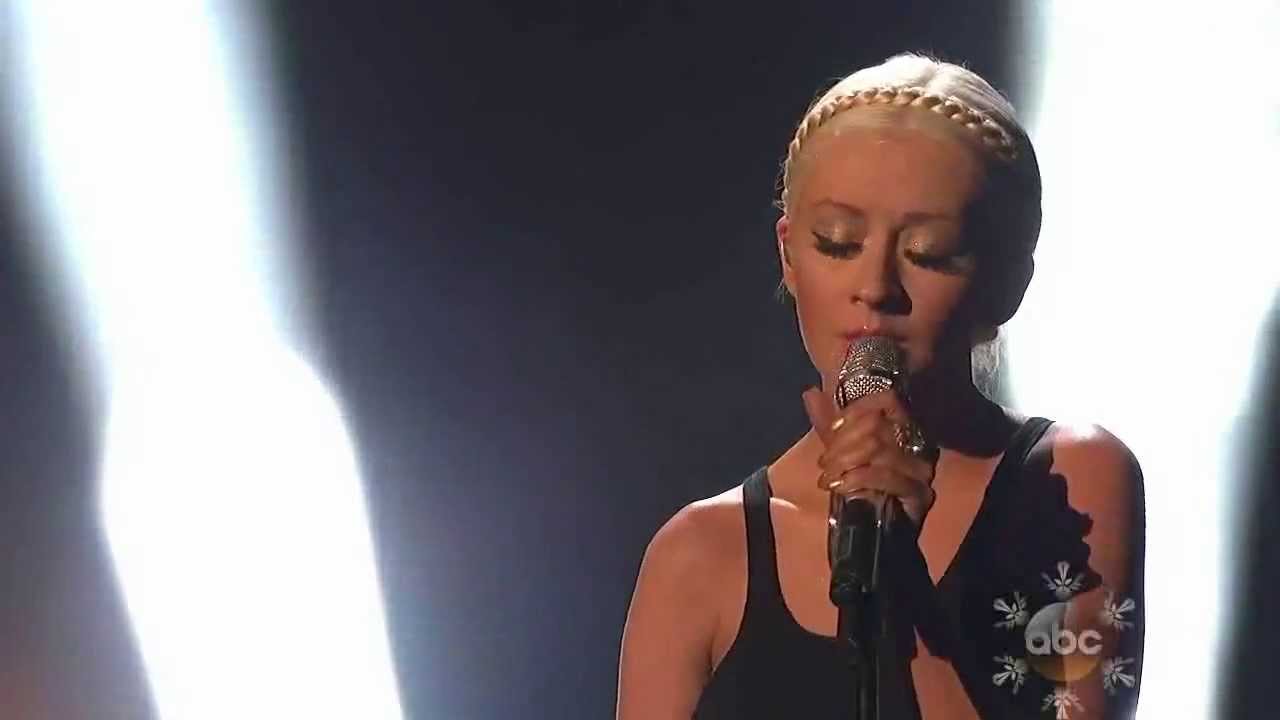 A woman with blonde hair styled in braids sings into a microphone, spotlighted on a dimly lit stage, during her emotive "Say Something" performance. Her expression is focused and intense.