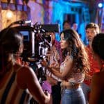 A woman adjusts a camera on a bustling film set, surrounded by actors and crew amidst colorful lighting, capturing the atmosphere of a dynamic movie production scene.