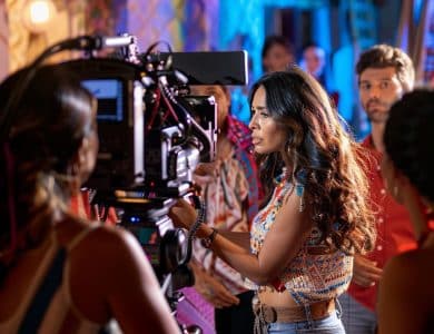 A woman adjusts a camera on a bustling film set, surrounded by actors and crew amidst colorful lighting, capturing the atmosphere of a dynamic movie production scene.