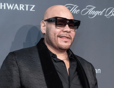 A man, reminiscent of Fat Joe, is wearing large black sunglasses, a black blazer with a velvet collar, and a black shirt. He is bald with a trimmed beard, smiling and standing in front of a dark gray backdrop with the words "Schwartz" and "The Angel Ball" partially visible.
