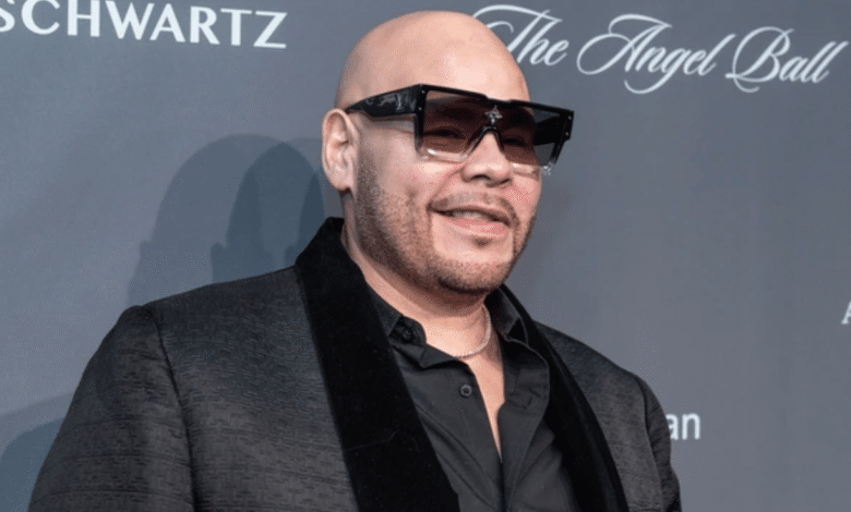 A man, reminiscent of Fat Joe, is wearing large black sunglasses, a black blazer with a velvet collar, and a black shirt. He is bald with a trimmed beard, smiling and standing in front of a dark gray backdrop with the words "Schwartz" and "The Angel Ball" partially visible.