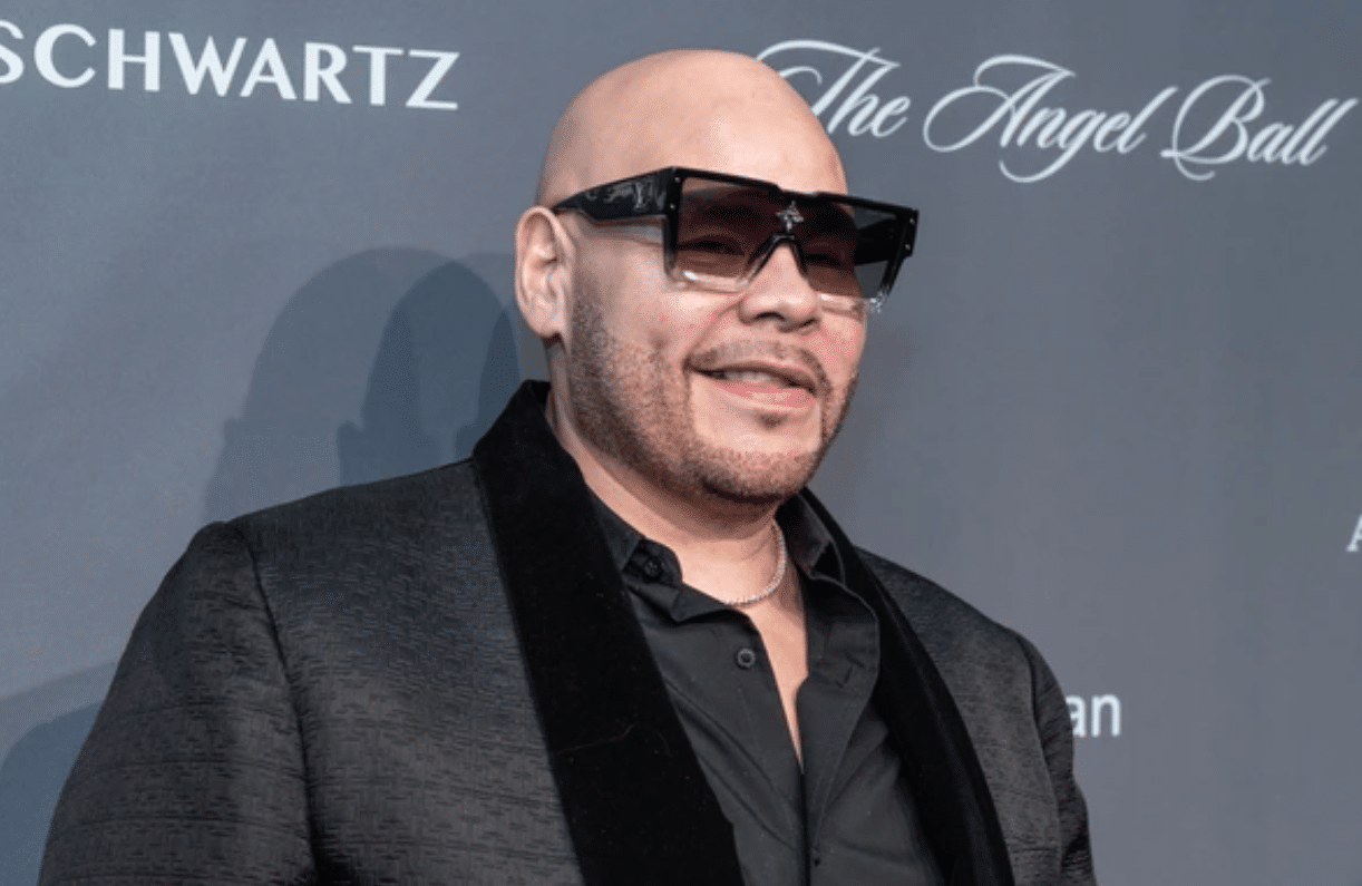A man, reminiscent of Fat Joe, is wearing large black sunglasses, a black blazer with a velvet collar, and a black shirt. He is bald with a trimmed beard, smiling and standing in front of a dark gray backdrop with the words "Schwartz" and "The Angel Ball" partially visible.