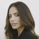 Geneis Rodriguez. A woman with long, wavy brown hair, parted in the middle, is shown against a plain white background. She is looking over her shoulder at the camera with a slight smile. Wearing a black top and exuding calm confidence, she evokes the poise of Genesis Rodriguez from Lioness Season 2.
