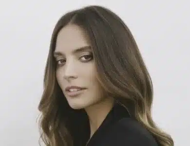 Geneis Rodriguez. A woman with long, wavy brown hair, parted in the middle, is shown against a plain white background. She is looking over her shoulder at the camera with a slight smile. Wearing a black top and exuding calm confidence, she evokes the poise of Genesis Rodriguez from Lioness Season 2.