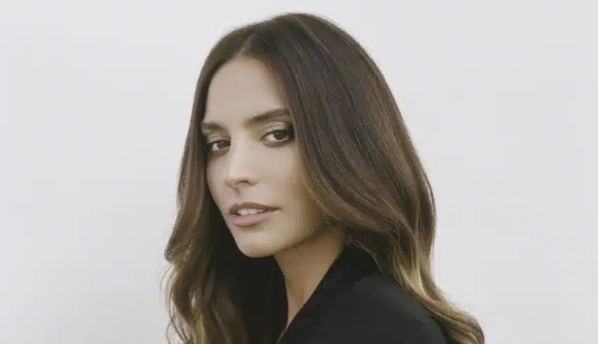 Geneis Rodriguez. A woman with long, wavy brown hair, parted in the middle, is shown against a plain white background. She is looking over her shoulder at the camera with a slight smile. Wearing a black top and exuding calm confidence, she evokes the poise of Genesis Rodriguez from Lioness Season 2.