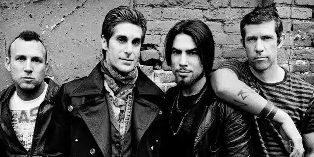 Janes Addiction, A black-and-white photo shows four men standing against a brick wall. The man on the left has short-cropped hair and a light-colored T-shirt. The second man wears a dark jacket and a patterned scarf. The third has long dark hair and a goatee. The fourth man wears a striped shirt with his arm resting on the third man's shoulder.