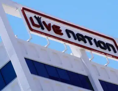 A photo of a white building with a large red and black "Live Nation" sign on top against a clear blue sky. The sign, mounted on the edge of the building and supported by metal brackets, hints at the ongoing Live Nation lawsuit discussions. The building has dark blue tinted windows on the upper floor.