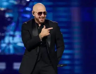 Pitbull, A bald man with a beard and sunglasses performs on stage wearing a black suit. He holds a microphone in his right hand and points with a smile using his left hand. The background is vibrant with blue and purple lighting, suggesting a lively concert atmosphere.