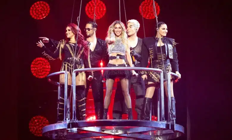 Five performers of the musical group RBD stand on a circular suspended platform, each dressed in unique, elaborate costumes. They are lit from above and behind by red stage lights arranged in a circular pattern. The central performer stands slightly ahead, all members smiling and engaging with the audience below.