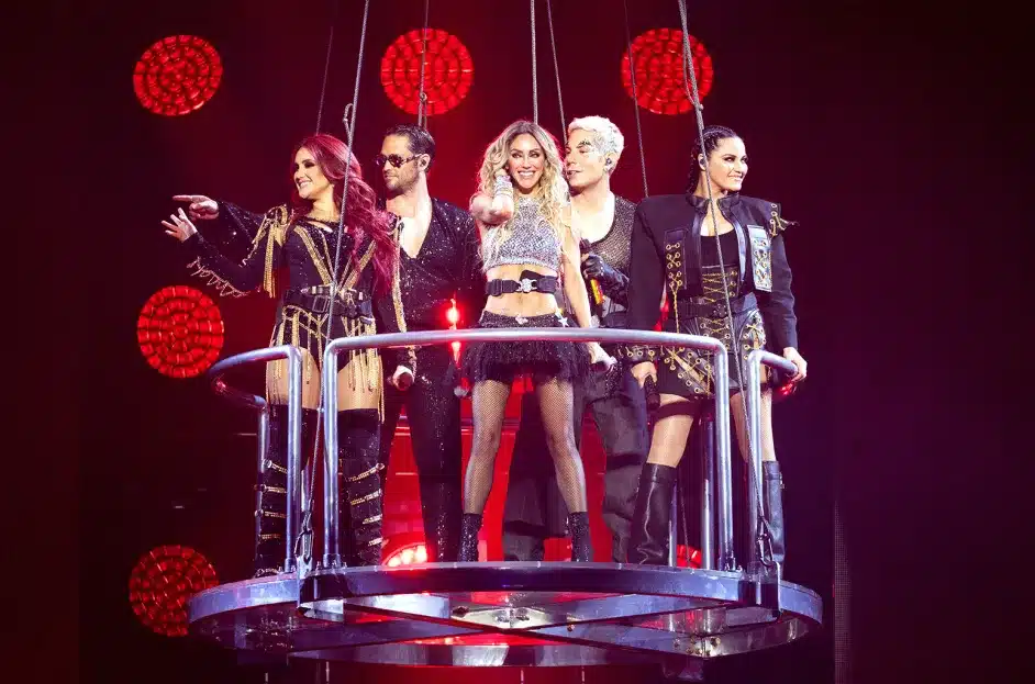 Five performers of the musical group RBD stand on a circular suspended platform, each dressed in unique, elaborate costumes. They are lit from above and behind by red stage lights arranged in a circular pattern. The central performer stands slightly ahead, all members smiling and engaging with the audience below.