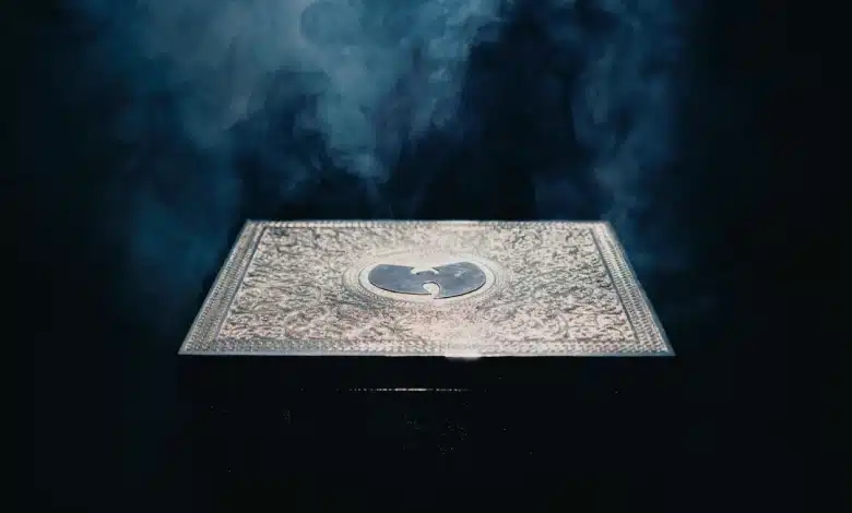 Wu-Tang Clan Once Upon a Time in Shaolin Alum, A square, ornately decorated silver box sits against a dark, smoky background. The box features an engraved emblem of a bird with outstretched wings, creating a glowing effect in the misty ambiance. The light emphasizes the intricate design of the box's surface.