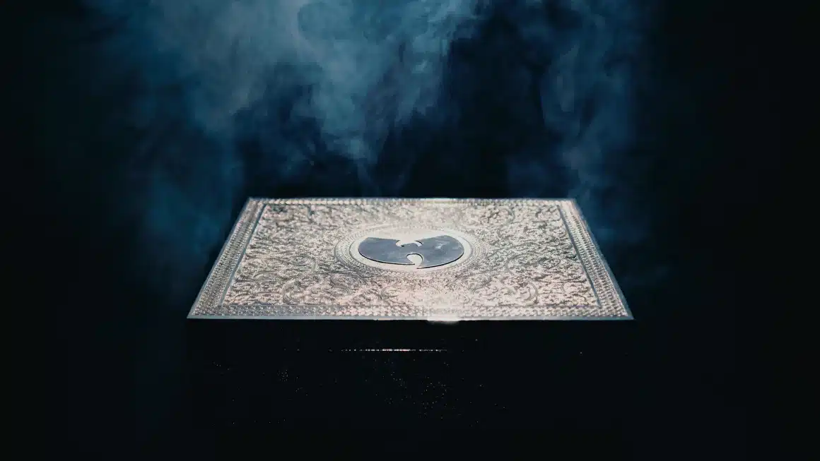 Wu-Tang Clan Once Upon a Time in Shaolin Alum, A square, ornately decorated silver box sits against a dark, smoky background. The box features an engraved emblem of a bird with outstretched wings, creating a glowing effect in the misty ambiance. The light emphasizes the intricate design of the box's surface.