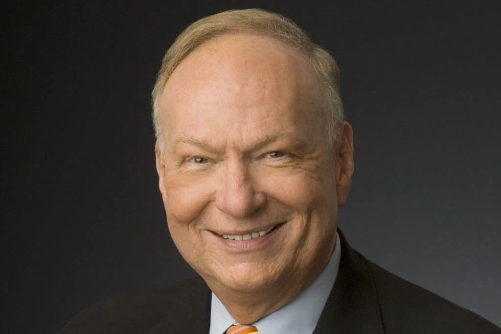 A portrait of an elderly caucasian man with a warm smile, wearing a dark suit, light blue shirt, and a blue tie. he has a clean-shaven face and short, light gray hair. the background is dark and plain.