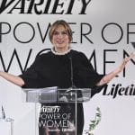 A woman stands at a podium with the "variety power of women" logo, smiling broadly with arms outstretched. she wears a black cape-like dress and a chunky pearl necklace.