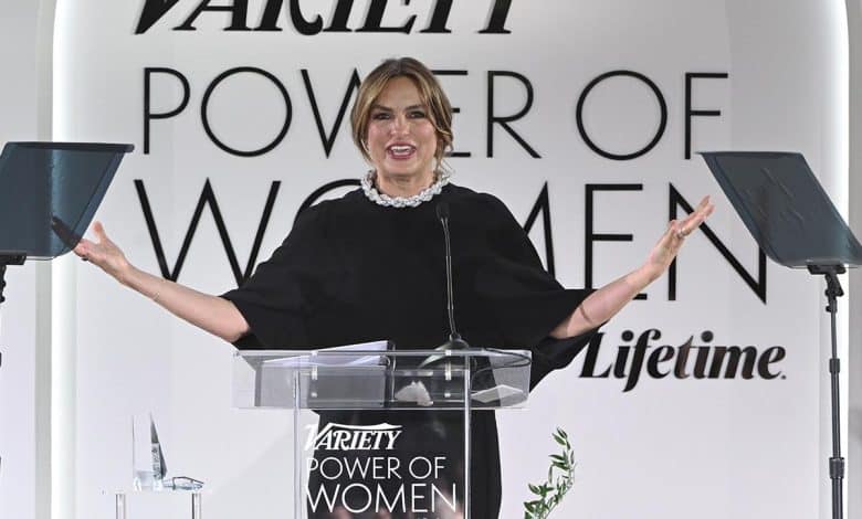 A woman stands at a podium with the "variety power of women" logo, smiling broadly with arms outstretched. she wears a black cape-like dress and a chunky pearl necklace.