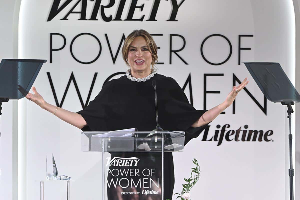 A woman stands at a podium with the "variety power of women" logo, smiling broadly with arms outstretched. she wears a black cape-like dress and a chunky pearl necklace.