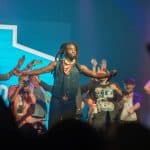 A dynamic concert scene showing a performer with braided hair and a scarf reaching out to an excited audience with raised hands, under blue stage lighting, with blurred fans and other performers in the background.