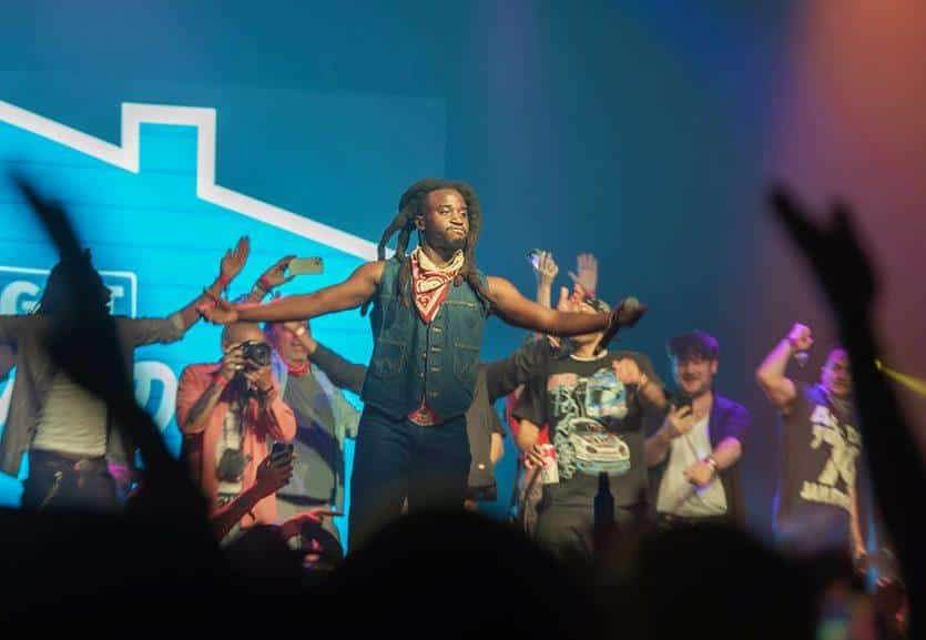 A dynamic concert scene showing a performer with braided hair and a scarf reaching out to an excited audience with raised hands, under blue stage lighting, with blurred fans and other performers in the background.
