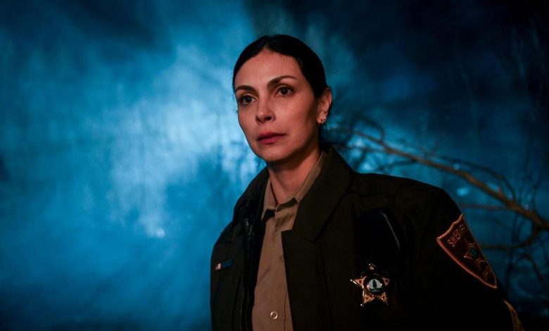 A close-up of a serious-looking female sheriff in a dark uniform, featuring a badge, against a moody, blue smoke-filled background with blurred branches. her face is lit warmly, contrasting with the cold background.