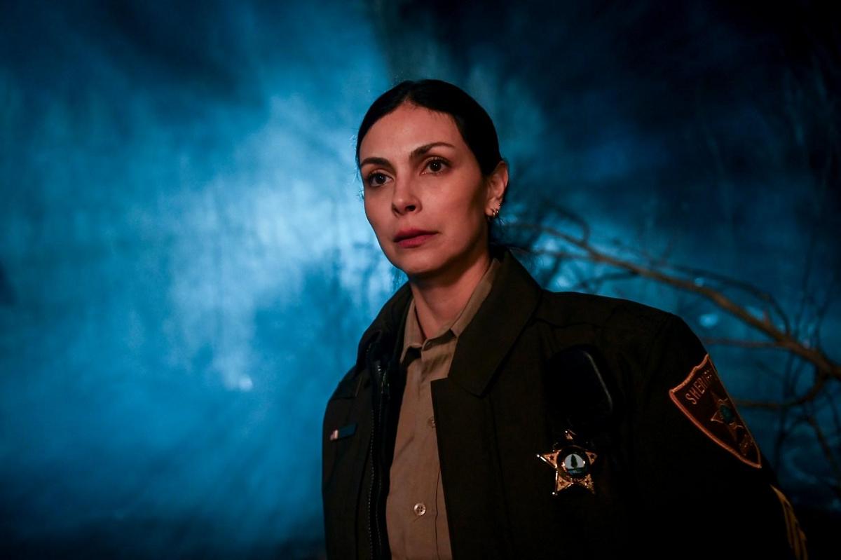A close-up of a serious-looking female sheriff in a dark uniform, featuring a badge, against a moody, blue smoke-filled background with blurred branches. her face is lit warmly, contrasting with the cold background.