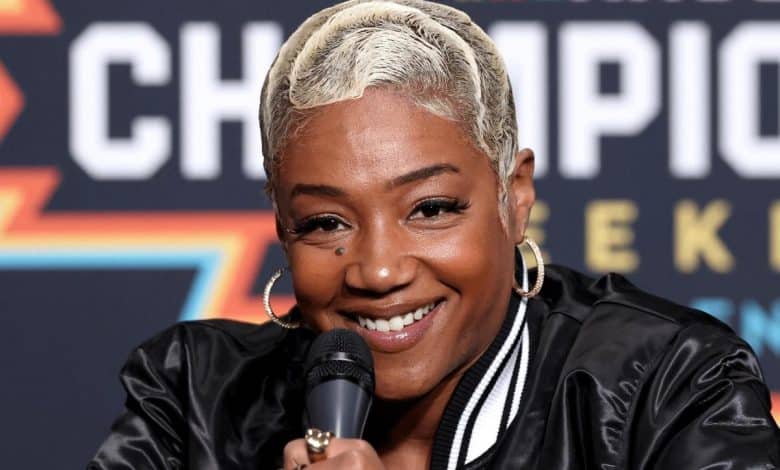 A woman with short platinum blonde hair smiles warmly while holding a microphone, sitting in front of a colorful backdrop featuring diagonal orange and blue stripes with text. she wears a black and white striped jacket and hoop earrings.