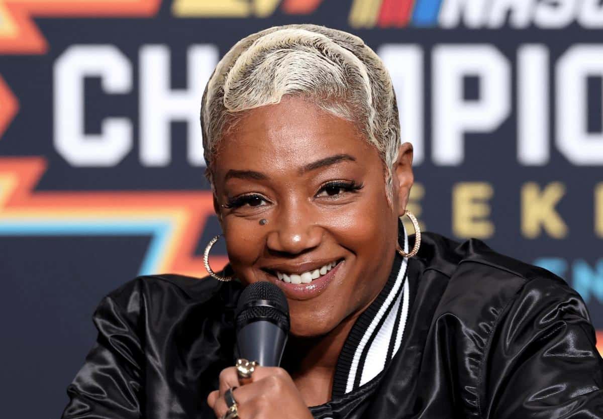 A woman with short platinum blonde hair smiles warmly while holding a microphone, sitting in front of a colorful backdrop featuring diagonal orange and blue stripes with text. she wears a black and white striped jacket and hoop earrings.