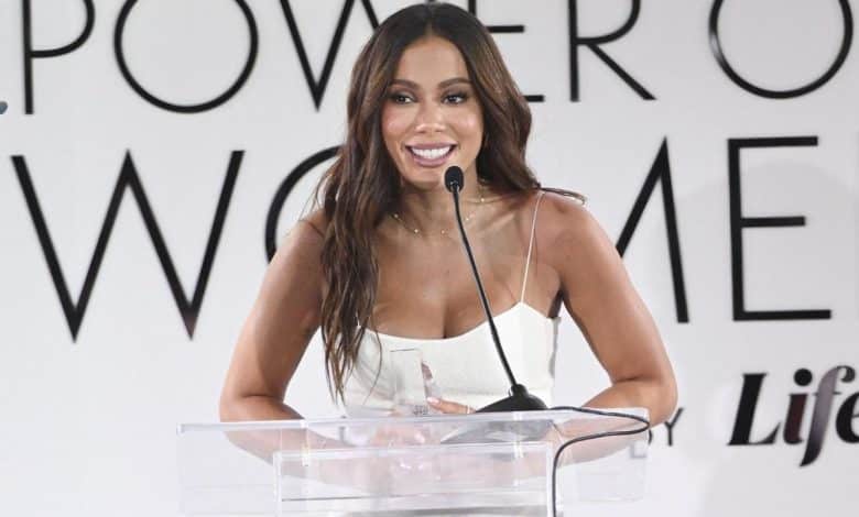 A woman speaking at a podium with the text "power of women" in the background. she is wearing a stylish white top and smiling warmly, her microphone positioned in front.