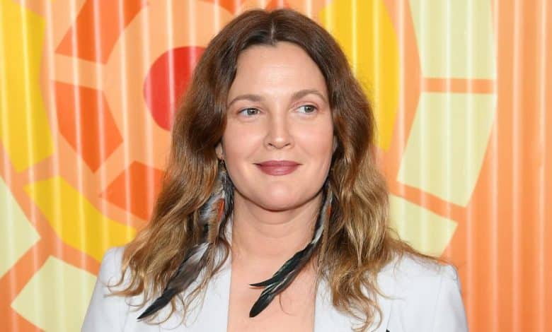 A woman with shoulder-length brown hair, wearing a light blazer and a black top, stands in front of a colorful abstract patterned background. she has a slight smile and is wearing feathered earrings.