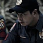 A focused male police officer wearing a dark uniform with embroidered badges gazes intently to the side. his expression is serious and contemplative, set against a blurred background of foliage.