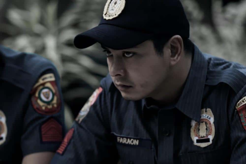 A focused male police officer wearing a dark uniform with embroidered badges gazes intently to the side. his expression is serious and contemplative, set against a blurred background of foliage.
