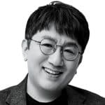A black and white portrait of an asian man smiling gently. he wears round glasses and a dark blazer over a light shirt. the background is plain, highlighting his cheerful expression.