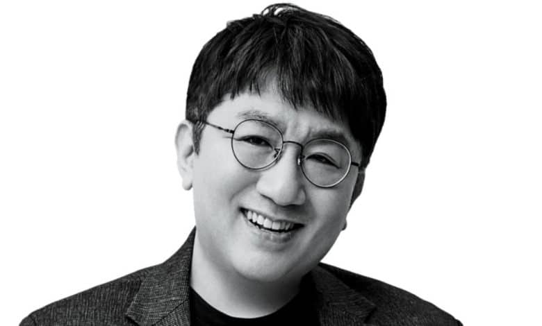 A black and white portrait of an asian man smiling gently. he wears round glasses and a dark blazer over a light shirt. the background is plain, highlighting his cheerful expression.