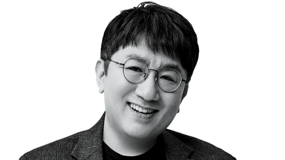 A black and white portrait of an asian man smiling gently. he wears round glasses and a dark blazer over a light shirt. the background is plain, highlighting his cheerful expression.