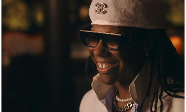 A close-up portrait of a joyful black woman wearing sunglasses, a white hat with a chanel logo, and pearl necklaces. she is smiling broadly in a dimly lit, warm-toned setting.