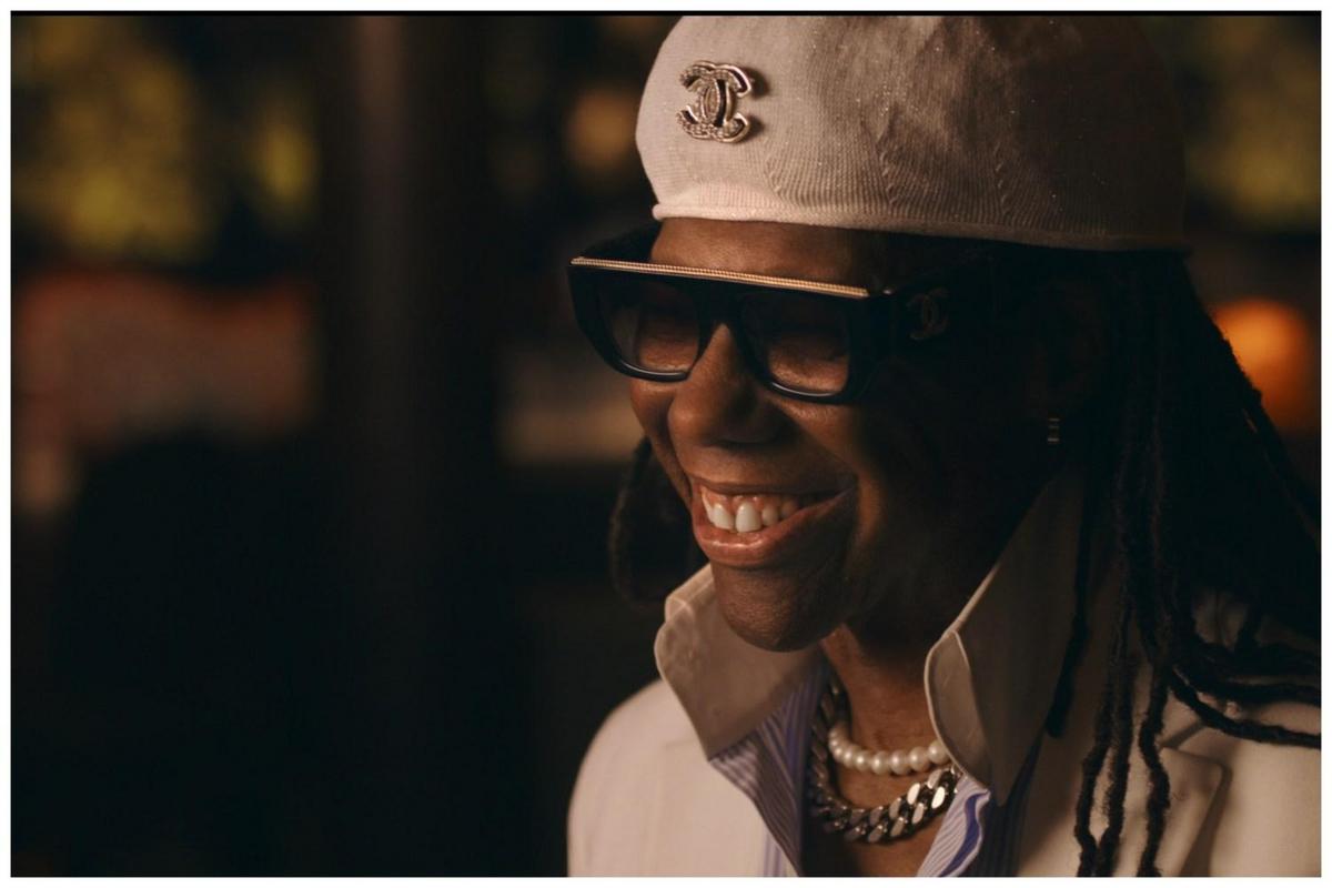 A close-up portrait of a joyful black woman wearing sunglasses, a white hat with a chanel logo, and pearl necklaces. she is smiling broadly in a dimly lit, warm-toned setting.